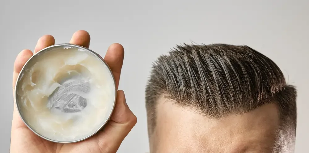 Person holding open container of hair pomade next to neatly styled hair.